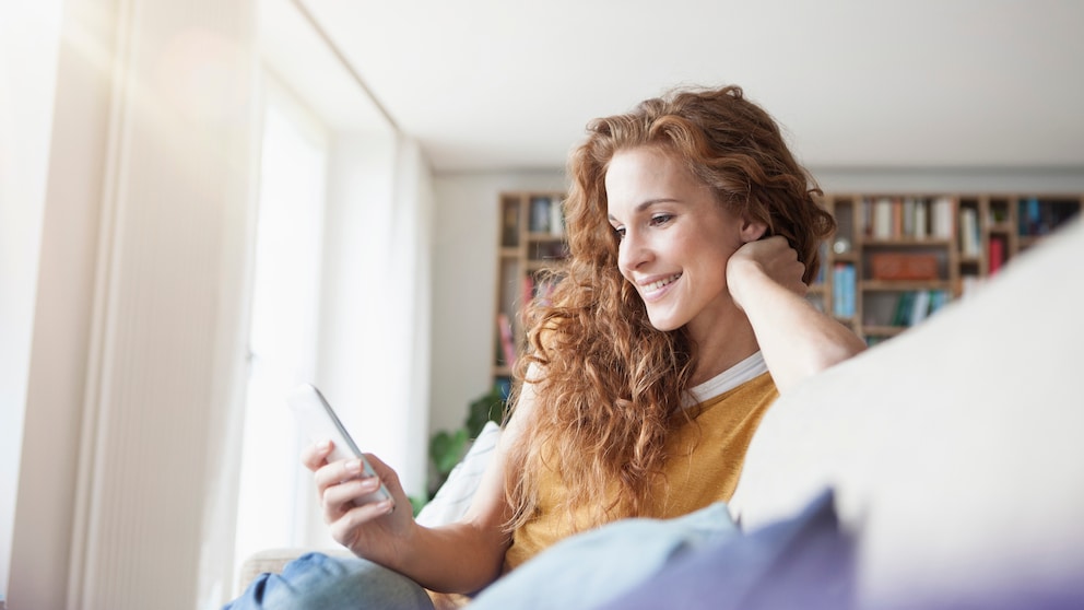 Frau grinst ins Handy auf dem Sofa. Es sei ihr zu wünschen, dass sie eine süße Nachricht bekommen hätte, anstatt online zu shoppen