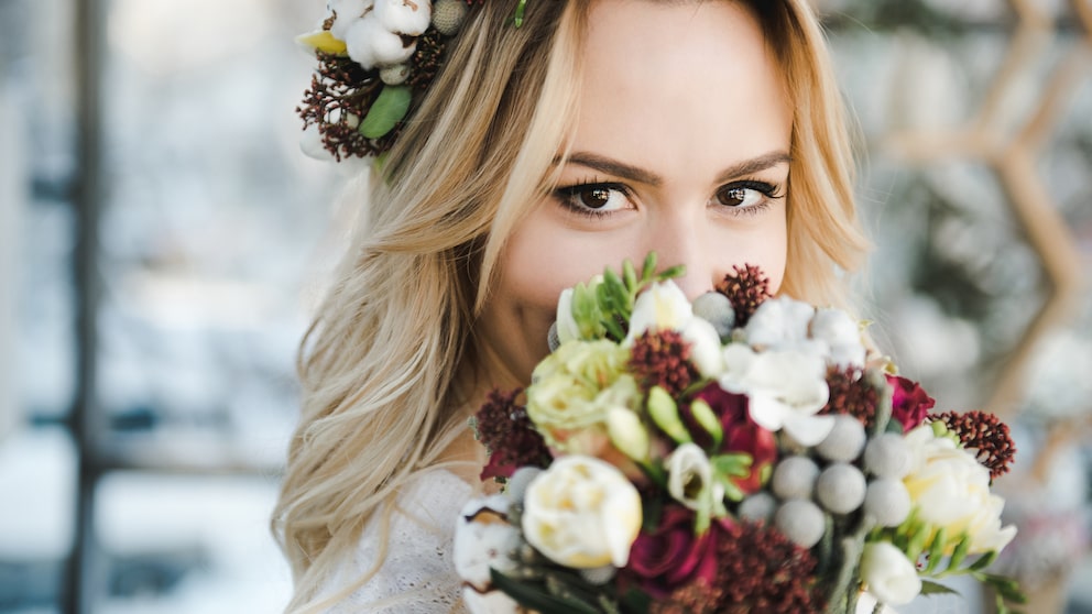 Frau mit Strauß und Brautkleid. Sie lächelt verschmitzt und wirkt, als würde sie ihren Namen nach der Hochzeit behalten. Bitte nicht persönlich nehmen, natürlich können Frauen, die ihren Namen behalten, aussehen, wie sie wollen. Es ist ja auch nur ein Beispielbild