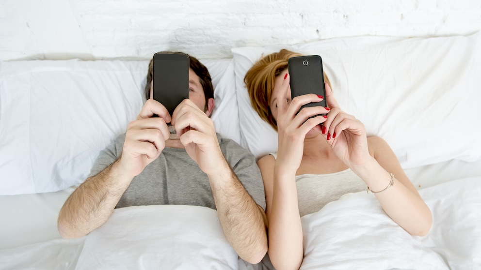 Ein Pärchen liegt im Bett. Beide halten ein Smartphone in den Händen