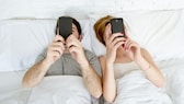 Ein Pärchen liegt im Bett. Beide halten ein Smartphone in den Händen