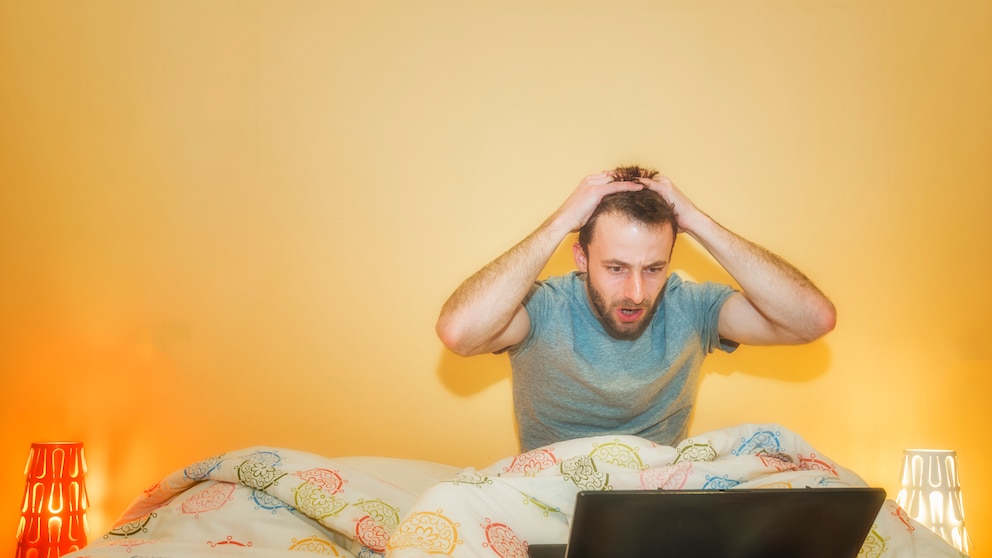 mann sitzt im Bett mit seinem Laptop und guckt geschockt