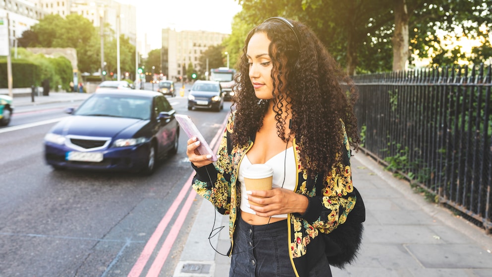 Frau trägt Kopfhörer und hält ihr Handy an einer Straße in der Hand