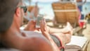 Ein Mann mit Basecap liegt auf Strandliege und schaut auf sein Smartphone