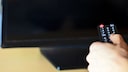 Ein Daumen einer Hand ist auf einer Fernbedienung vor einem TV zu sehen