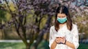 Frau mit Maske steht im Park und schaut aufs Smartphone