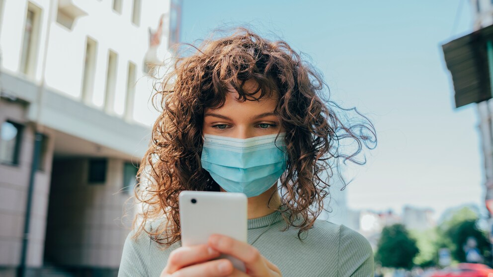 Frau mit Mund-Nasen-Maske schaut aufs Smartphone