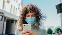 Frau mit Mund-Nasen-Maske schaut aufs Smartphone