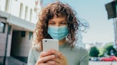 Frau mit Mund-Nasen-Maske schaut aufs Smartphone