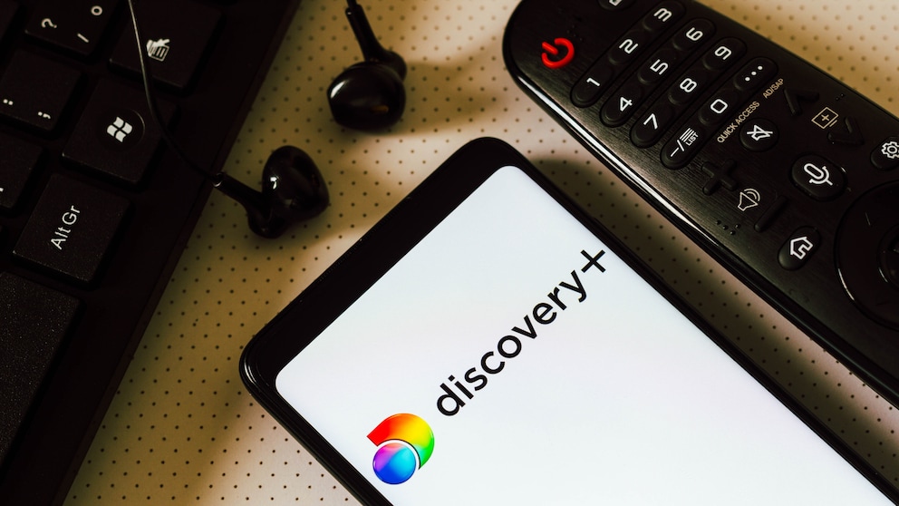 Discovery+ Logo auf Smartphone neben einer Fernbedienung