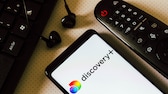 Discovery+ Logo auf Smartphone neben einer Fernbedienung