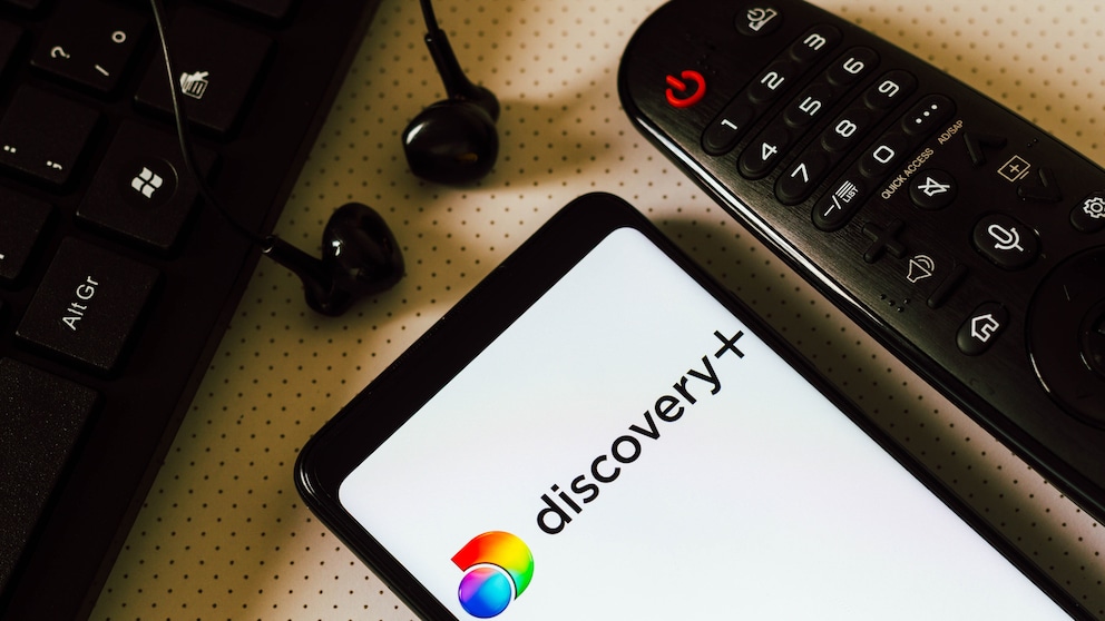 Discovery+ Logo auf Smartphone neben einer Fernbedienung