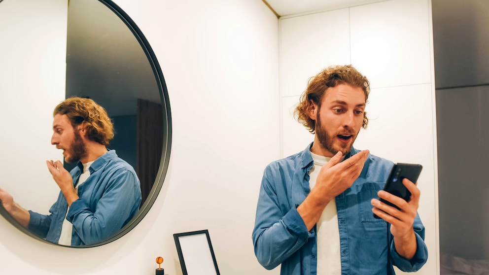 Junger Mann mit Smartphone im Bad ist geschockt weil er beim Dating mit einem Nacktbild erpresst wird – Sextortion