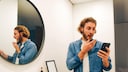 Junger Mann mit Smartphone im Bad ist geschockt weil er beim Dating mit einem Nacktbild erpresst wird – Sextortion