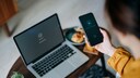 Junge Frau hält Smartphone mit Virenschutz an einem Schreibtisch, auf dem ein MacBook steht.