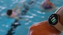 Smart Watch eines Schwimmmeisters im Vordergrund, dahinter unscharf das Schwimmbecken