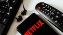 Netflix Account Sharing Gebühr Folgen: Netflix Logo auf Smartphone neben Fernbedienung und Computertatstatur