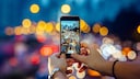 Symbolbild: Frau fotografiert mit Smartphone den abendlichen Verkehr, der mit Bokeh-Effekt im Hintergrund zu sehen ist.