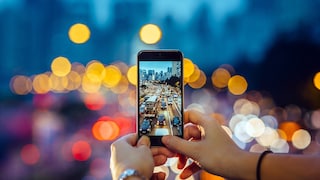 Symbolbild: Frau fotografiert mit Smartphone den abendlichen Verkehr, der mit Bokeh-Effekt im Hintergrund zu sehen ist.