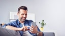 Mann sitzt mit einem Smartphone in der Hand auf einem Sofa.