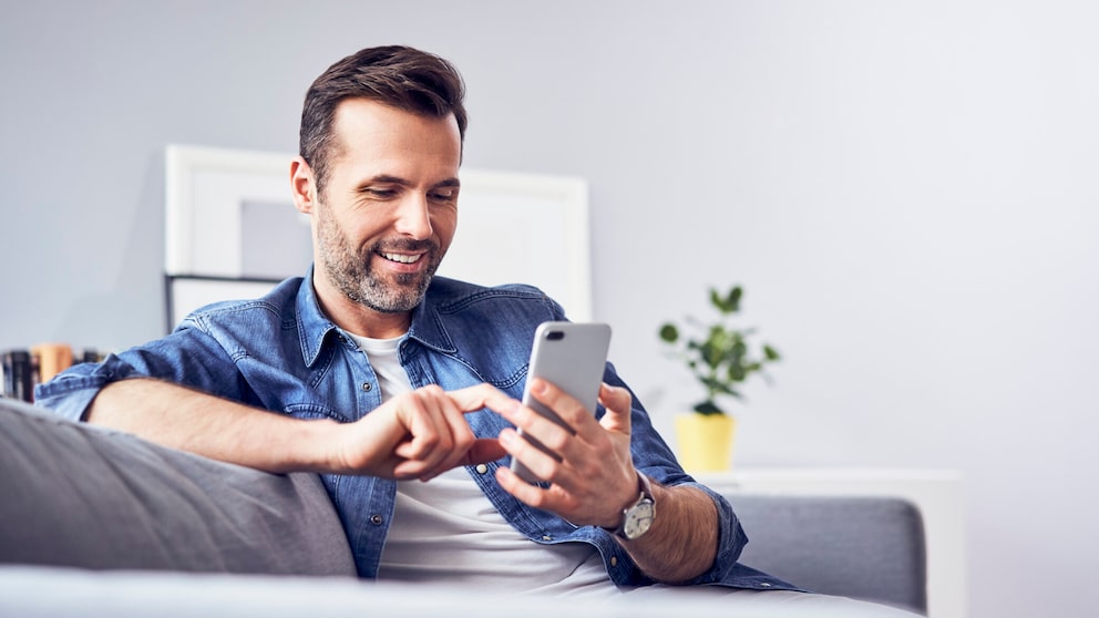 Mann sitzt mit einem Smartphone in der Hand auf einem Sofa.