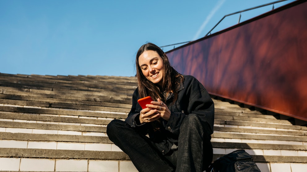 Symbolbild: junge Frau sitzt draußen und beschäftigt sich mit ihrem Smartphone