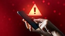 Malware Symbolbild: Hand mit Smartphone, dahinter ein großes Warnsignal