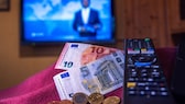 Die Summe von 18,94 Euro liegt auf einem Tisch neben der Fernbedienung für einen Fernseher. Der Rundfunkbeitrag soll ab 2025 vorläufigen Berechnungen von Finanzexperten zufolge auf 18,94 Euro steigen. Das geht nach Informationen der Deutschen Presse-Agentur aus einem am Freitag bekanntgewordenen Entwurf des Expertengremiums KEF hervor.