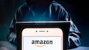Mit Kapuze vermummte Person sitzt an einem Laptop, im Vordergrund ein Handy-Display mit Amazon-Logo.