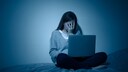 Frau sitzt mit vorgeschlagener Hand mit Laptop auf dem Scho&szlig; auf dem Bett.