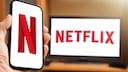 Person hält Handy mit Netflix-Logo in Hand, dahinter TV mit Schriftzug des Streamers.