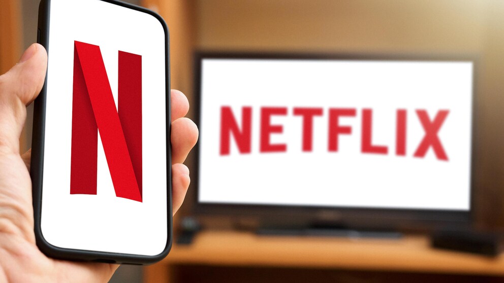 Person hält Handy mit Netflix-Logo in Hand, dahinter TV mit Schriftzug des Streamers.