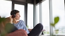 Symbolbild: Frau hört mit Overear-Kopfhörern Musik und schaut aus dem Fenster