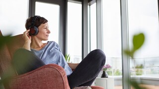 Symbolbild: Frau hört mit Overear-Kopfhörern Musik und schaut aus dem Fenster