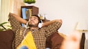 Symbolbild: junger Mann hört mit Kopfhörer auf dem Sofa ein Hörbuch