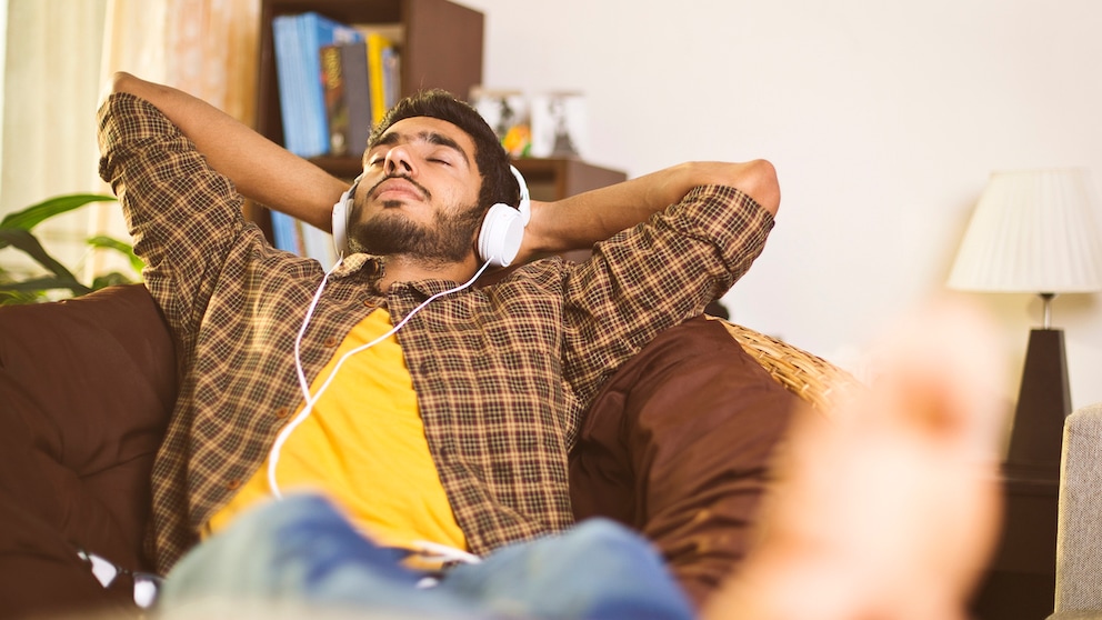 Symbolbild: junger Mann hört mit Kopfhörer auf dem Sofa ein Hörbuch