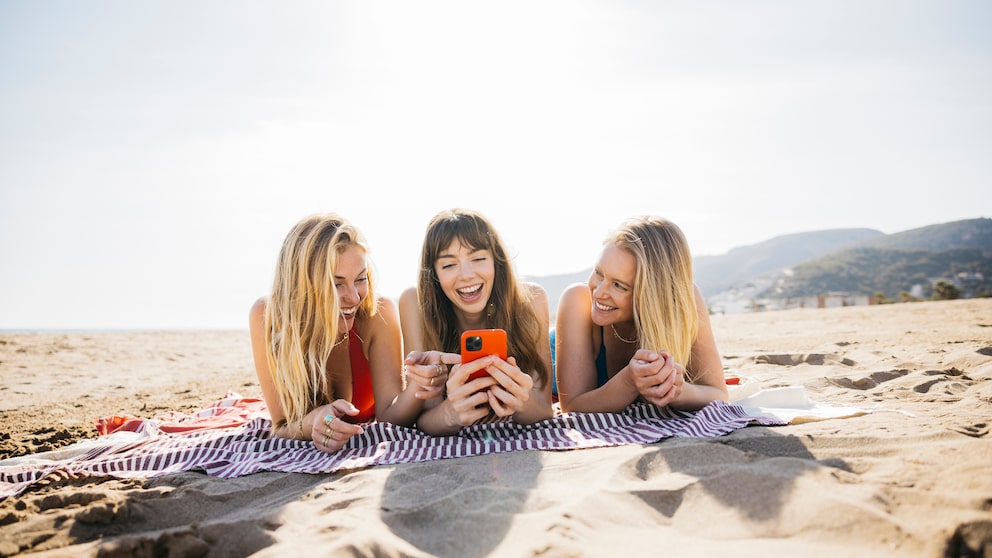 Drei Frauen liegen am Strand und schauen auf ein Handy