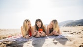 Drei Frauen liegen am Strand und schauen auf ein Handy