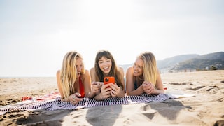 Drei Frauen liegen am Strand und schauen auf ein Handy