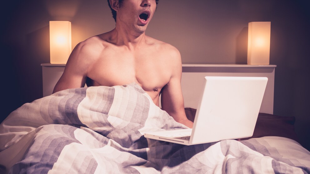 Mann sitzt im Bett mit aufgeklapptem Laptop