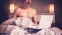Mann sitzt im Bett mit aufgeklapptem Laptop