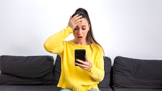 Frau sitzt mit Smartphone in der Hand auf dem Sofa und schlägt sich erschrocken die Hand an den Kopf