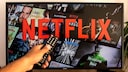 Person zeigt mit einer Fernbedienung auf einen Fernseher, auf dem Netflix zu sehen ist
