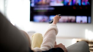 Person sitzt auf dem Sofa und zeigt mit der Fernbedienung auf den Fernseher