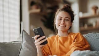 Frau sitzt lächelnd mit ihrem Smartphone auf dem Sofa