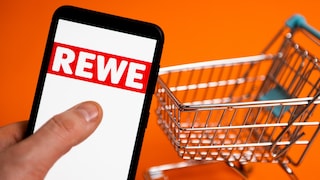 Rewe-Logo auf einem Smartphone, daneben ein Einkaufswagen