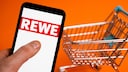 Rewe-Logo auf einem Smartphone, daneben ein Einkaufswagen