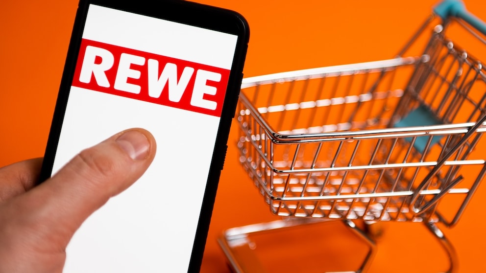 Rewe-Logo auf einem Smartphone, daneben ein Einkaufswagen