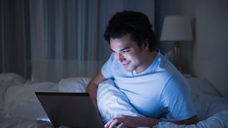 Mann schaut im Bett lächelnd auf seinen Laptop