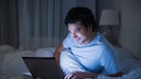 Mann schaut im Bett lächelnd auf seinen Laptop