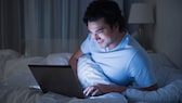 Mann schaut im Bett lächelnd auf seinen Laptop